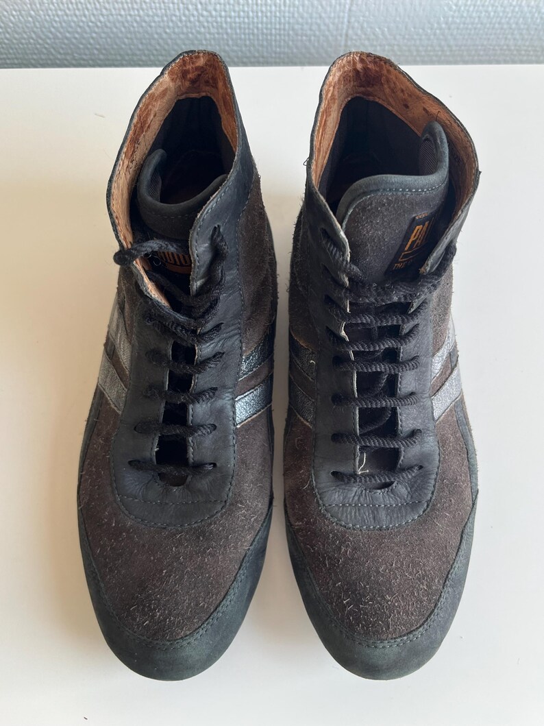 Pu&ograve; includere: Un paio di sneakers alte in pelle scamosciata marrone e pelle nera. Le sneakers hanno lacci neri, una striscia argentata e una suola nera. L'interno &egrave; di colore marrone chiaro.