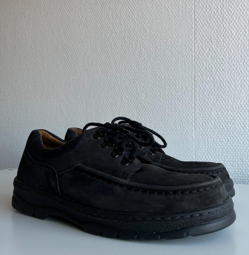 May include: A pair of black suede shoes with thick soles. The shoes have a classic lace-up design with black laces and a rounded toe. The shoes are displayed on a white surface against a textured white wall.