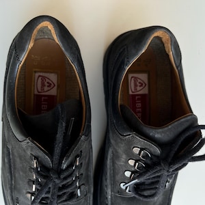 May include: A pair of black leather shoes with laces, metal eyelets, and a textured appearance. The shoes have a low-profile design with a padded collar and a visible brand label inside. The soles are not visible.