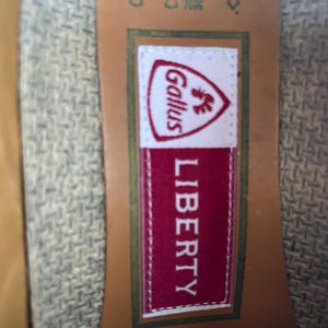 May include: Close-up of a label with the text "LIBERTY" in white on a red background. Above the text is a white label with the word "Gallus" and a rooster emblem. The label is on a textured fabric.