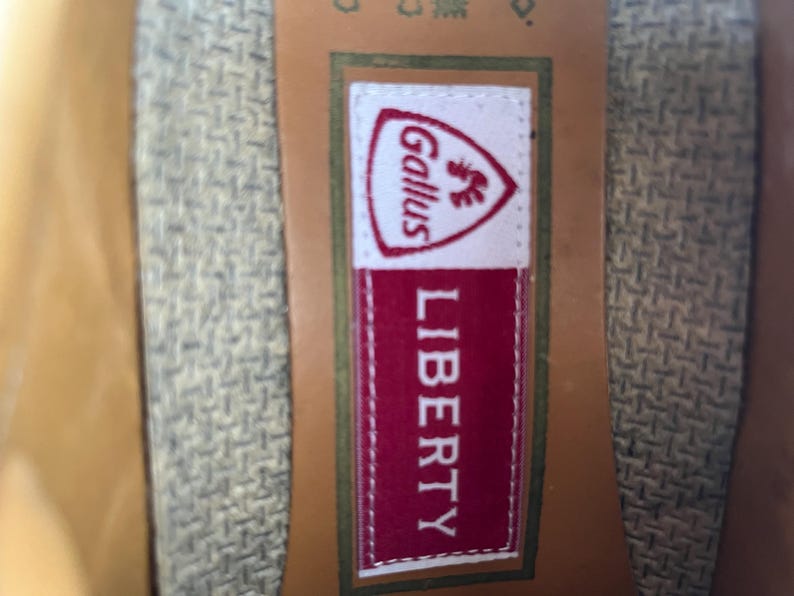 May include: Close-up of a label with the text "LIBERTY" in white on a red background. Above the text is a white label with the word "Gallus" and a rooster emblem. The label is on a textured fabric.