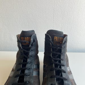 Pu&ograve; includere: Paio di sneakers alte in pelle scamosciata marrone e pelle nera. Le sneakers presentano lacci neri, un colletto imbottito e il marchio "PALLADIUM" in lettere dorate. Strisce argentate accentuano i lati.