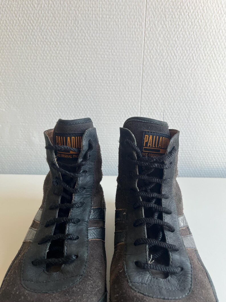 Pu&ograve; includere: Paio di sneakers alte in pelle scamosciata marrone e pelle nera. Le sneakers presentano lacci neri, un colletto imbottito e il marchio "PALLADIUM" in lettere dorate. Strisce argentate accentuano i lati.