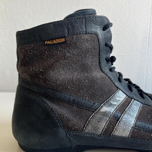 Pu&ograve; includere: Primo piano di una sneaker alta. La scarpa &egrave; principalmente nera e marrone, con strisce argentate sul lato. Il nome del marchio "PALLADIUM" &egrave; visibile in lettere arancioni. La scarpa ha lacci neri e una texture simile alla pelle scamosciata.