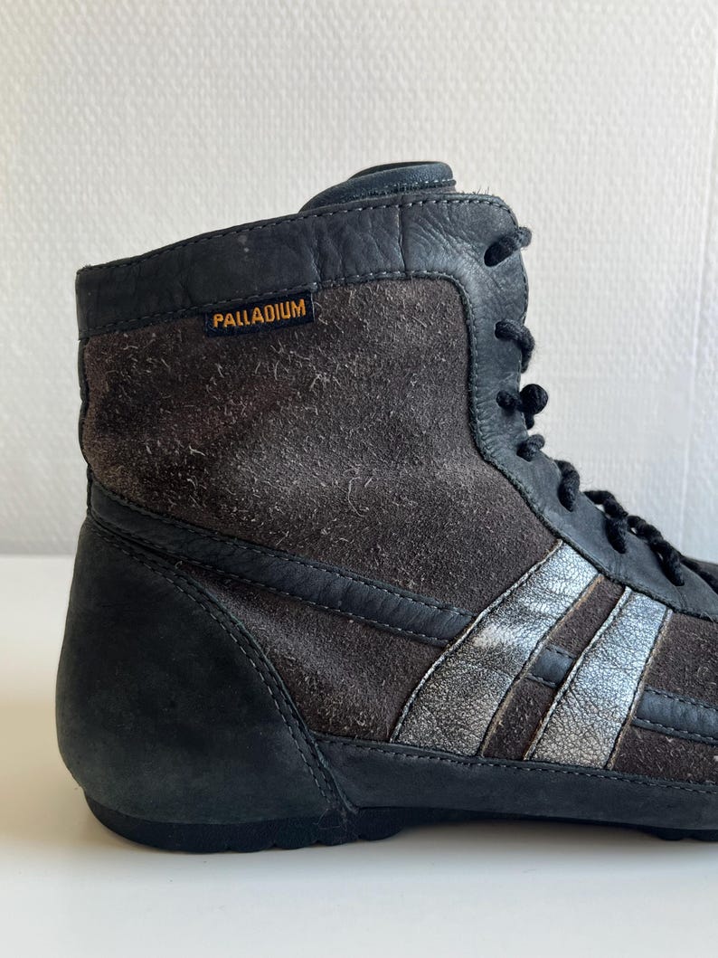 Pu&ograve; includere: Primo piano di una sneaker alta. La scarpa &egrave; principalmente nera e marrone, con strisce argentate sul lato. Il nome del marchio "PALLADIUM" &egrave; visibile in lettere arancioni. La scarpa ha lacci neri e una texture simile alla pelle scamosciata.
