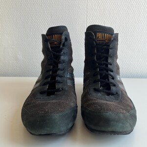 Pu&ograve; includere: Paio di sneakers alte nere e marroni con lacci neri. Le sneakers hanno una consistenza simile alla pelle scamosciata e la parola "PALLADIUM" &egrave; stampata in arancione sulla linguetta. Le scarpe sono su una superficie bianca.