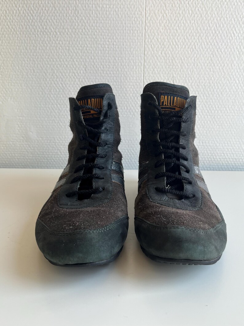 Pu&ograve; includere: Paio di sneakers alte nere e marroni con lacci neri. Le sneakers hanno una consistenza simile alla pelle scamosciata e la parola "PALLADIUM" &egrave; stampata in arancione sulla linguetta. Le scarpe sono su una superficie bianca.