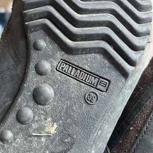 Pu&ograve; includere: Primo piano della suola di una scarpa grigio scuro. La suola presenta un motivo strutturato con il marchio "PALLADIUM" e la taglia "39" stampati. La suola della scarpa &egrave; realizzata in un materiale resistente.