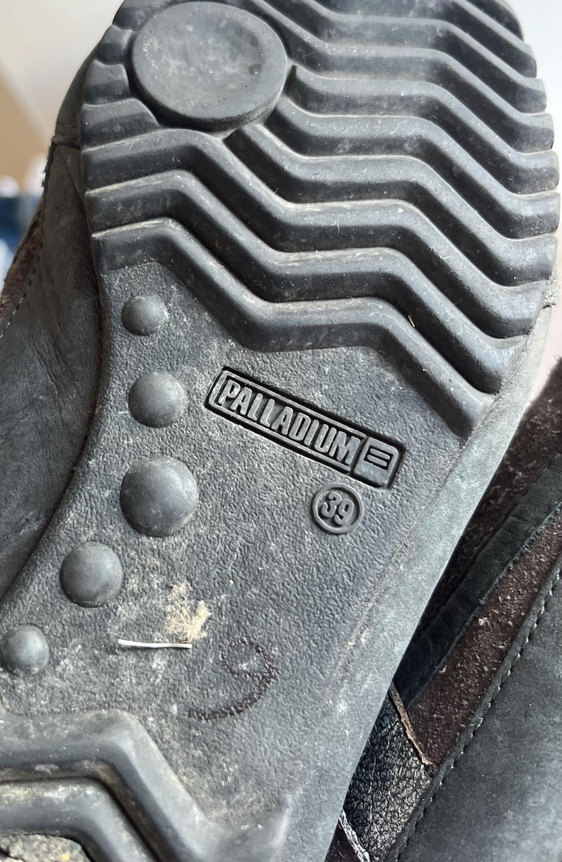 Pu&ograve; includere: Primo piano della suola di una scarpa grigio scuro. La suola presenta un motivo strutturato con il marchio "PALLADIUM" e la taglia "39" stampati. La suola della scarpa &egrave; realizzata in un materiale resistente.