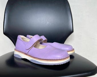 May include: A pair of lavender Mary Jane style shoes with a strap closure. The shoes have a white sole with a light brown and gray trim. The shoes are sitting on a black chair.