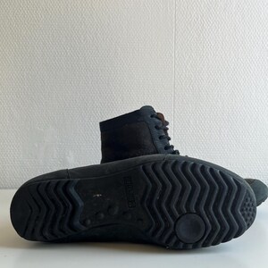 Pu&ograve; includere: Stivali in pelle nera con design stringato. Gli stivali hanno una suola strutturata per l'aderenza e un gambale di media altezza. L'immagine mostra la suola di uno stivale e il lato dell'altro, entrambi su una superficie bianca.