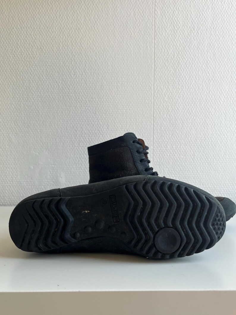 Pu&ograve; includere: Stivali in pelle nera con design stringato. Gli stivali hanno una suola strutturata per l'aderenza e un gambale di media altezza. L'immagine mostra la suola di uno stivale e il lato dell'altro, entrambi su una superficie bianca.