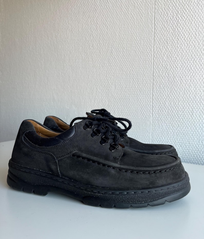 May include: A pair of black suede shoes with thick soles and black laces. The shoes have a classic, casual design, suitable for everyday wear. The shoes are displayed on a white surface.