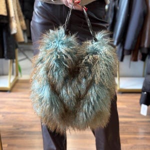 May include: A teal and brown fluffy handbag with silver handles. The bag has a shaggy texture and a unique design. The bag is held by a person in brown trousers.
