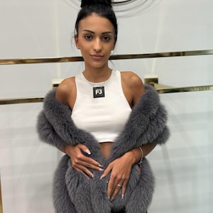 May include: A grey fur stole worn over a white vest top. The stole is made of soft, fluffy material. A silver necklace with a square pendant is visible. The person is wearing black trousers.