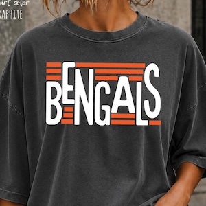 May include: A graphite-coloured t-shirt featuring the word BENGALS in white capital letters. The text is layered over horizontal orange stripes. The shirt has a crew neckline and short sleeves, with the text t-shirt colour GRAPHITE printed above.