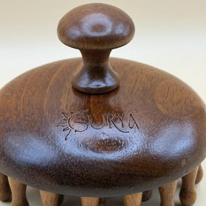 May include: A wooden massage tool with a mushroom-shaped handle and multiple rounded legs. The tool is dark brown with the word "SURYA" engraved on the top. The massage tool is designed for body massage.