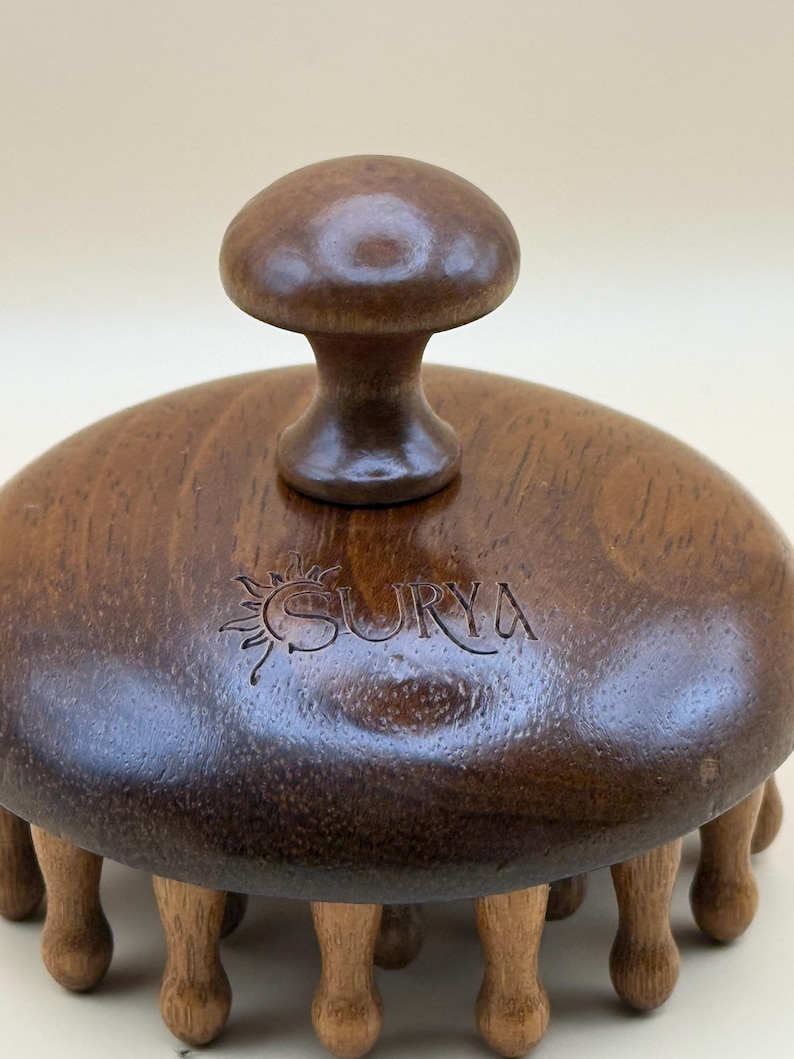 May include: A wooden massage tool with a mushroom-shaped handle and multiple rounded legs. The tool is dark brown with the word "SURYA" engraved on the top. The massage tool is designed for body massage.
