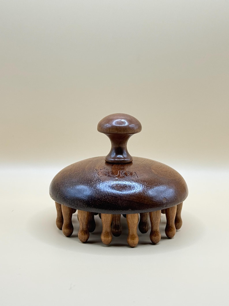 May include: A wooden massage tool with a dark brown, domed head and a small handle. The head has several small wooden legs extending downward. The word "SURYA" is etched on the top. The tool is designed for massage and relaxation.