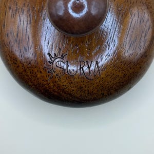 May include: Close-up of a wooden lid with a dark brown knob. The lid has a circular shape with a rich, textured grain. The word "SURYA" is engraved on the surface, accompanied by a sun-like emblem. The overall aesthetic is rustic and natural.