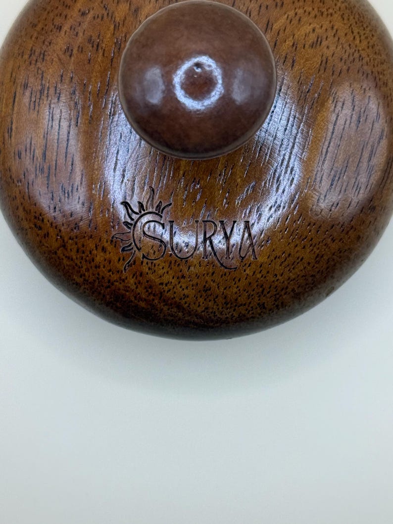 May include: Close-up of a wooden lid with a dark brown knob. The lid has a circular shape with a rich, textured grain. The word "SURYA" is engraved on the surface, accompanied by a sun-like emblem. The overall aesthetic is rustic and natural.
