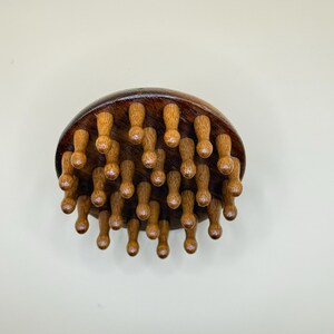 May include: A wooden massage tool with a dark brown, rounded top and numerous small, light brown pegs extending downward. The pegs are evenly spaced and appear to be designed for pressure point massage. The tool is set against a neutral background.