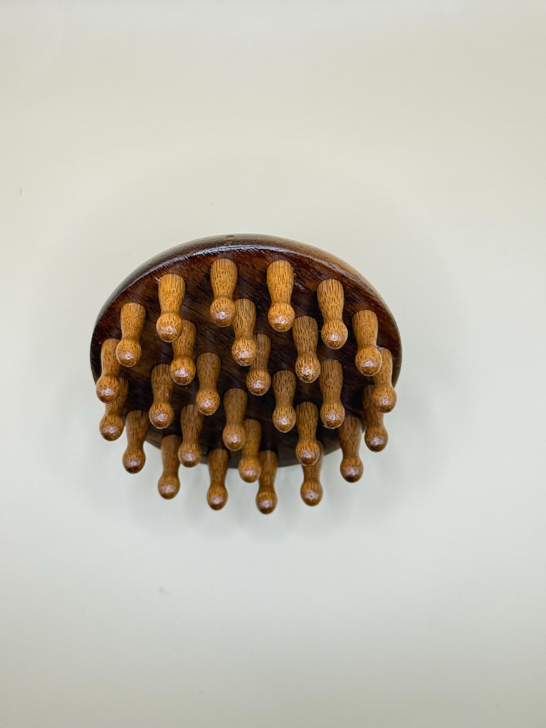 May include: A wooden massage tool with a dark brown, rounded top and numerous small, light brown pegs extending downward. The pegs are evenly spaced and appear to be designed for pressure point massage. The tool is set against a neutral background.