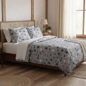 Op de afbeelding: Een grijze beddenset met een pootafdrukpatroon. De set bevat een dekbedovertrek en twee kussenslopen. De pootafdrukken zijn zwart en wit, en het dekbedovertrek ligt op een houten bedframe. Een beige deken ligt aan het voeteneinde van het bed.