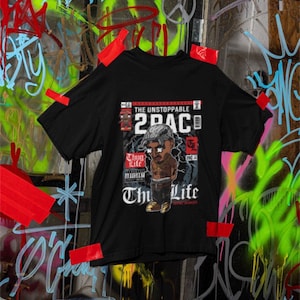 May include: Black t-shirt featuring a cartoon illustration of 2Pac with the text "The Unstoppable 2Pac" and "Thug Life". The shirt is taped to a wall covered in graffiti art.