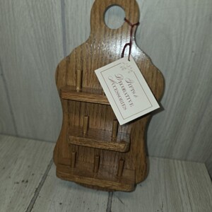May include: A wall-mounted wooden rack with three horizontal shelves and six vertical pegs for storage. The light brown oak-toned wood features a cutout handle at the top. A rectangular white tag is attached with string, reading Gifts & DECORATIVE ACCESSORIES.