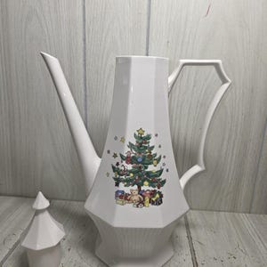 May include: A white porcelain coffee pot with a faceted body and a matching lid. The front features a colorful illustration of a decorated Christmas tree with ornaments, a star topper, and wrapped gifts with a teddy bear at the base.