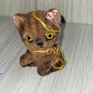 May include: A vintage brown cat figurine with yellow eyes and a black nose. The cat is adorned with yellow and pink thread accents. The figurine has a textured, flocked appearance and is posed in a seated position.