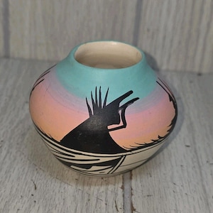 May include: A small, round ceramic pot with a wide opening. The pot features a painted design with a gradient of colors, including pink, blue, and white. A black silhouette of a figure and geometric patterns are also present.