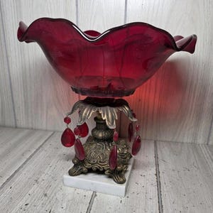 May include: A deep red glass bowl with a ruffled rim, supported by an ornate bronze-toned metal base featuring hanging red teardrop crystals. The base rests on a square white marble plinth, creating a decorative centerpiece with metallic accents.