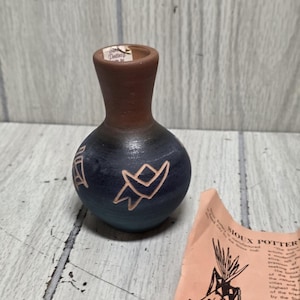 May include: A small, round, dark blue and brown clay vase with a narrow neck. The body features etched geometric patterns. A small paper label on the rim reads Sioux Pottery. Beside the vase is a light peach-colored paper with black text and a drawing.