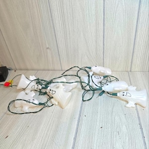 May include: A string of white plastic ghost-shaped lights with black facial features. The lights are connected by a green wire and are on a light-colored wooden surface. The lights are for Halloween.