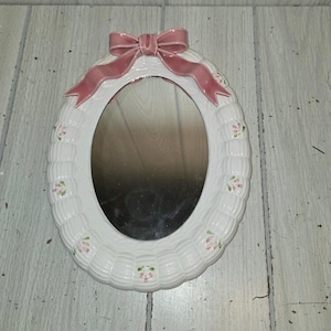 May include: Oval white ceramic mirror with a pink bow at the top. The frame has a woven texture with small pink and green floral accents. The mirror is a decorative piece.
