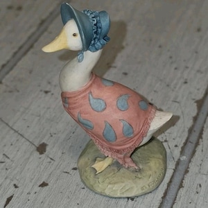 May include: A ceramic figurine of a white duck wearing a blue bonnet and a pink shawl with a blue paisley pattern. The duck stands on a textured, light green circular base. The base features the embossed text "BY SCOTLAND" near the edge.