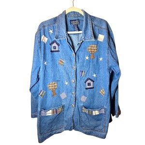 May include: A blue denim button-down jacket featuring embroidered appliques of houses, trees, stars, and plaid fabric patches. The jacket has a collar, long sleeves, and two large front pockets with plaid trim. A label reads Willow Ridge.