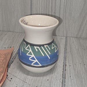 May include: A small, off-white ceramic vase with a flared rim and a rounded body. The midsection features horizontal bands of dark green and blue, decorated with white geometric line patterns. A small paper tag is attached to the inner rim.