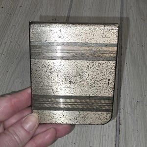 May include: A rectangular, vintage metal cigarette case with a silver finish. The case has horizontal bands of textured silver and a slightly worn appearance, suggesting age and use. The case is held in a hand.