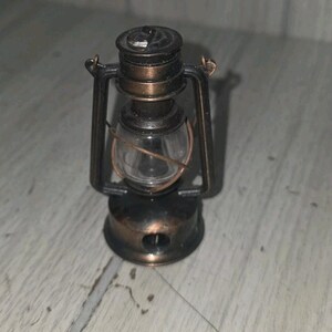 May include: A miniature oil lantern featuring a dark bronze-toned metal frame with a weathered finish. The lantern includes a clear glass globe at the center, a circular base with a small opening, and a top handle attached to the side support arms.