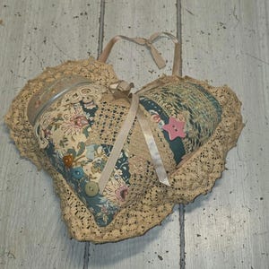 May include: A heart-shaped fabric ornament featuring a patchwork design of floral and solid teal fabric, edged with beige crochet lace. It is decorated with a pink star-shaped button, several round buttons, and a beige ribbon tied in a bow.