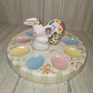 May include: A ceramic Easter egg holder with a white bunny figurine in the center. The bunny has floral accents. The holder is round with pastel-colored egg-shaped indentations and a green and white checkered border.