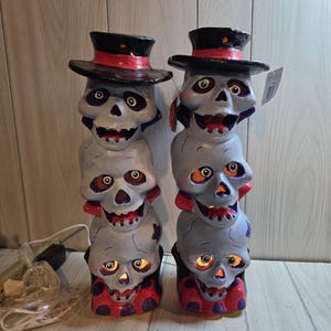 May include: Two vertical stacks of three light gray skulls, each wearing a black top hat with a red band. The skulls have green eyes and red-painted teeth, with internal lighting visible through the eye sockets and mouths. A white power cord is attached.