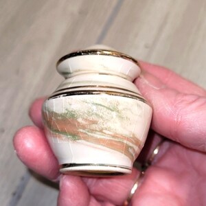 May include: A small, rounded ceramic vessel featuring a cream-colored base with marbled streaks of sage green and terracotta brown. The piece has a glossy finish with visible fine crackle patterns and is accented by thin, metallic gold trim.