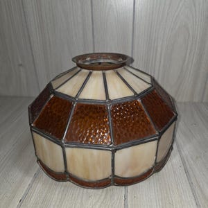 May include: A vintage lamp shade with a geometric design. The shade features alternating panels of textured amber and cream-colored glass, framed by a metal border. The top has a metal ring for attachment.