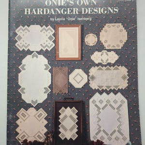 May include: A softcover book titled ONIE'S OWN HARDANGER DESIGNS by Leona Onie Isenberg, compiled by Susan L. Meier and Rosalyn K. Watnemo. The cover features a dark blue floral background with various white and beige Hardanger embroidery patterns.
