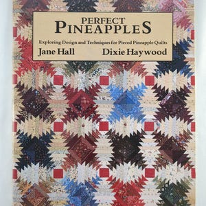 Puede incluir: Un libro titulado "Perfect Pineapples" con un colorido patrón de edredón en la portada. La portada presenta una pancarta beige con el título en letras negras grandes. Debajo del título está el subtítulo "Exploring Design and Techniques for Pieced Pineapple Quilts".