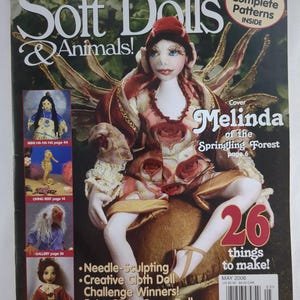 Puede incluir: La portada de la revista "Soft Dolls & Animals!" presenta una muñeca con sombrero rojo y vestido floral. La portada incluye texto sobre patrones, escultura con aguja y cosas para hacer. También aparece el texto "Melinda of the Springling Forest".
